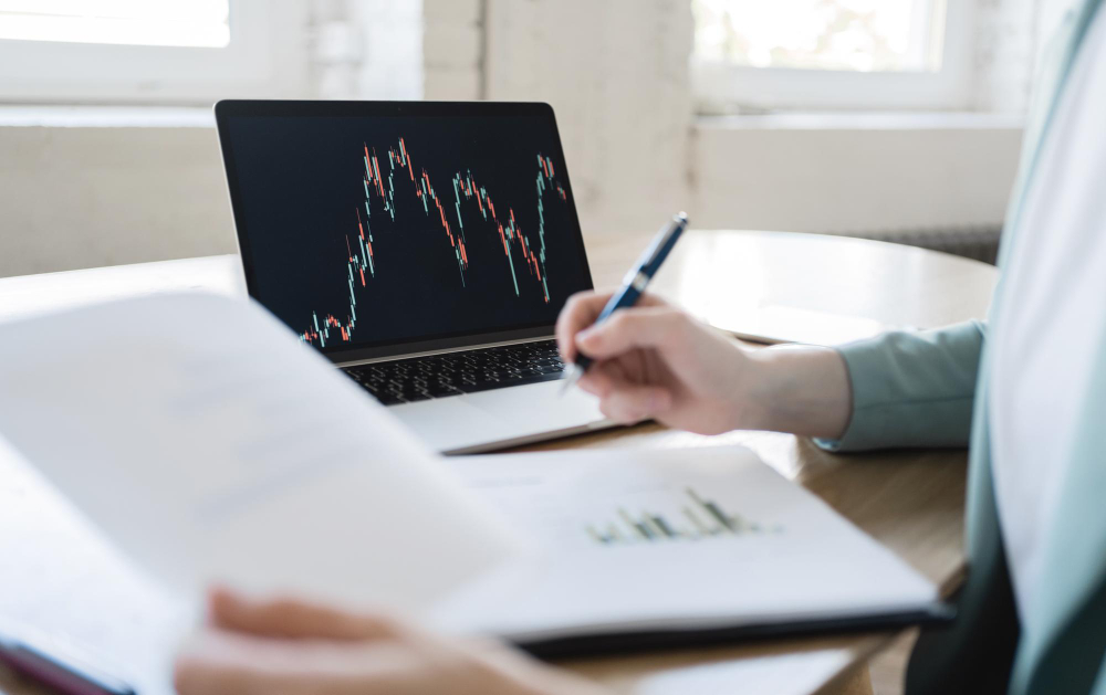 A person is studying financial charts on a laptop screen while holding a pen and reviewing a document, suggesting an analysis of market trends or investment research.
